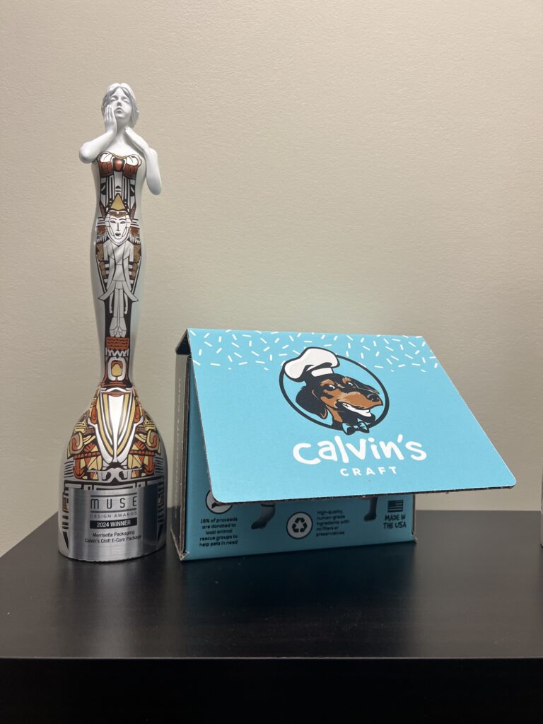 A Muse Design Award 2024 Winner trophy designed to look like a woman holding her right hand to her cheek and her left hand to her neck is colorful, silver-based with red, gold and vermillion accents. It stands next to a Calvin's Craft retail packaging box that is sky blue and features the face of a Dachshund dog wearing a Chef's hat and the word's Calvin's Craft underneath him. 