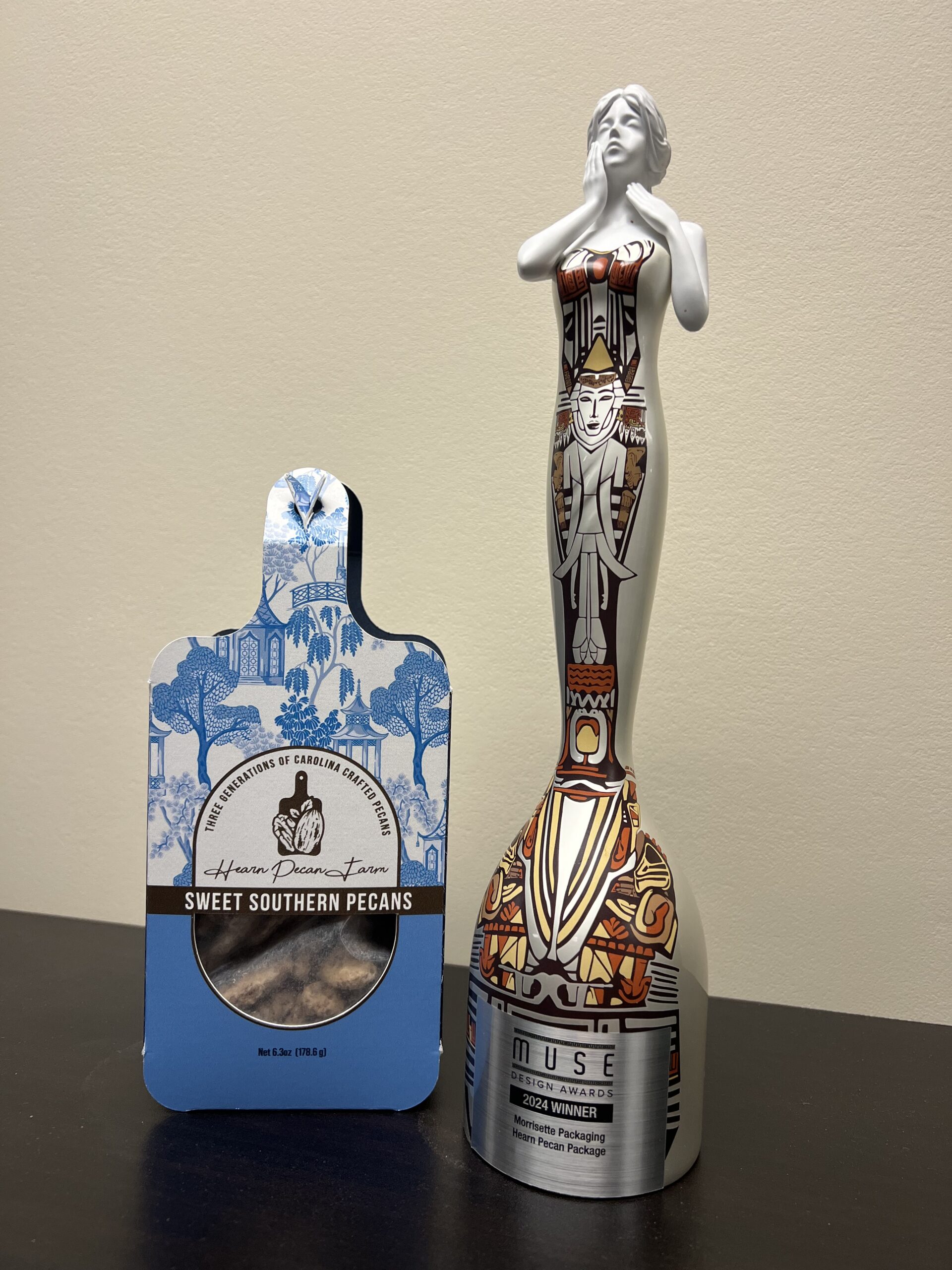 A Muse Design Award 2024 Winner trophy designed to look like a woman holding her right hand to her cheek and her left hand to her neck is colorful, silver-based with red, gold and vermillion accents. It stands next to a Hearn Pecan Farm retail packaging box that is blue and mirrors the company's logo which resembles a serving board.
